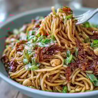 A steaming bowl of chili crisp noodles, glossy and coated in spicy, garlicky chili oil, topped with fresh scallions and crunchy peanuts.
